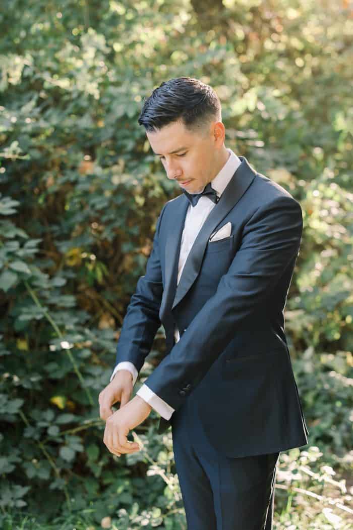 Elegant man in a tailored suit adjusting his cuff in a lush outdoor setting.
