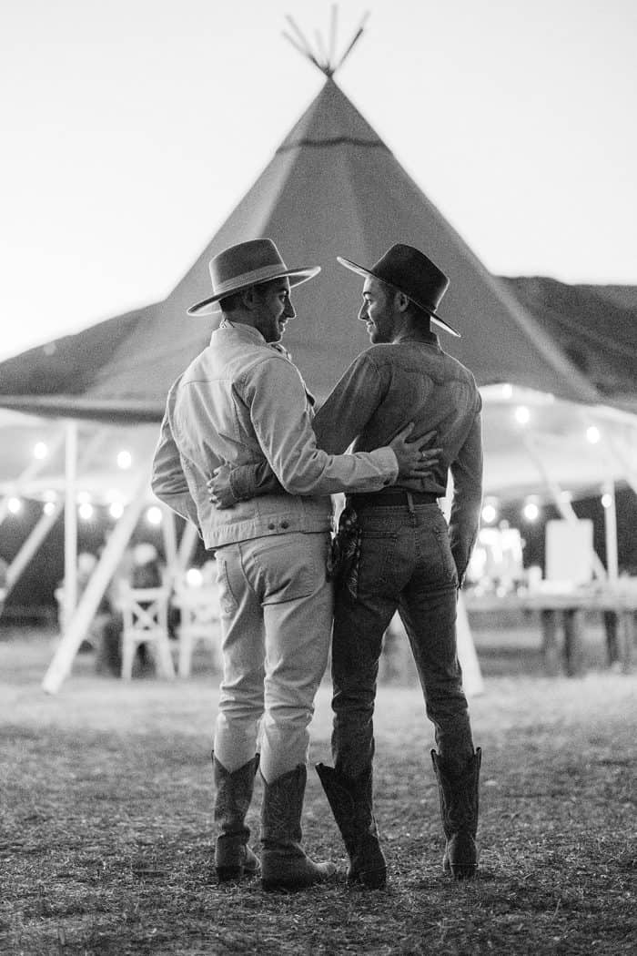 Two friends in cowboy hats embrace at outdoor festival with tipi in background.