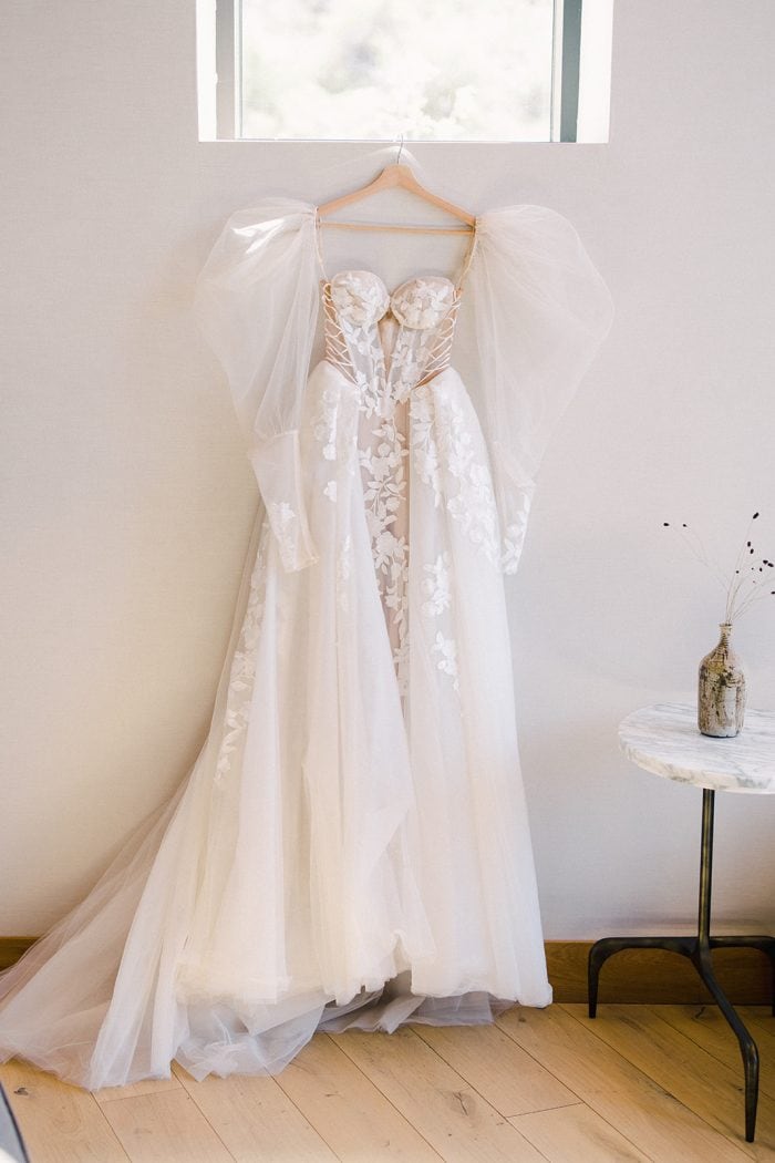 Wedding dress hanging on a wall hanger with tulle sleeves and lace details.