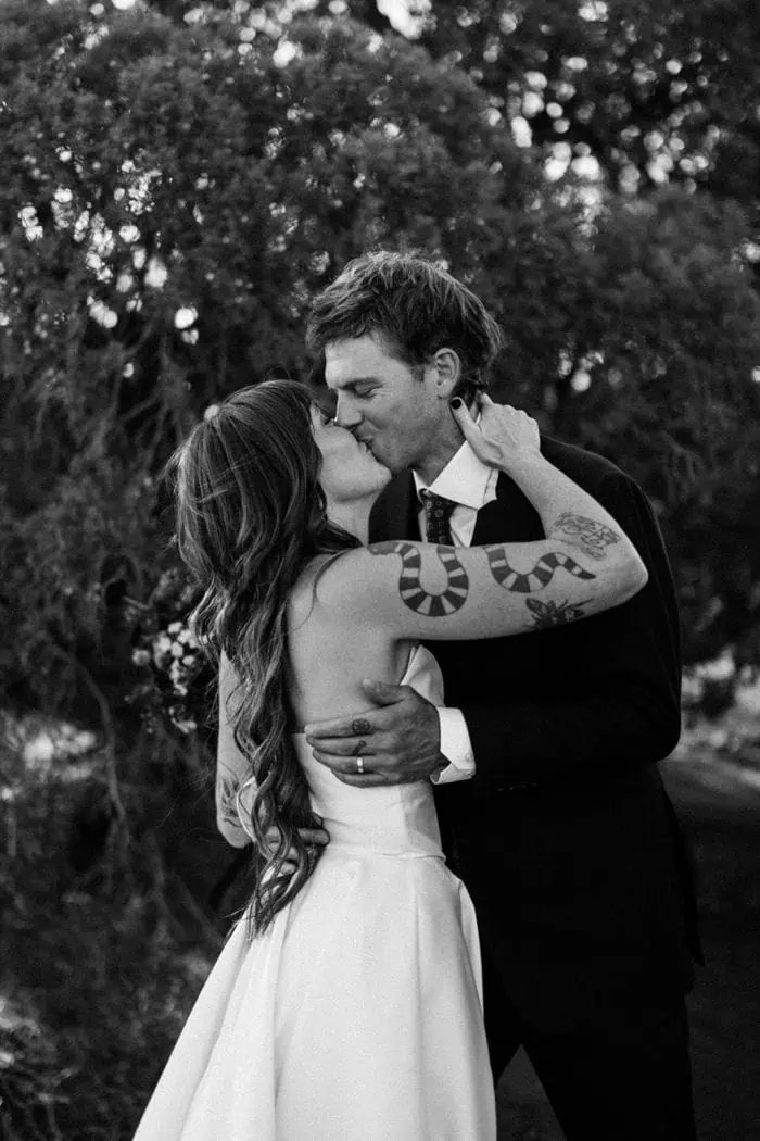 Kissing couple in wedding attire, embracing outdoors during sunset.
