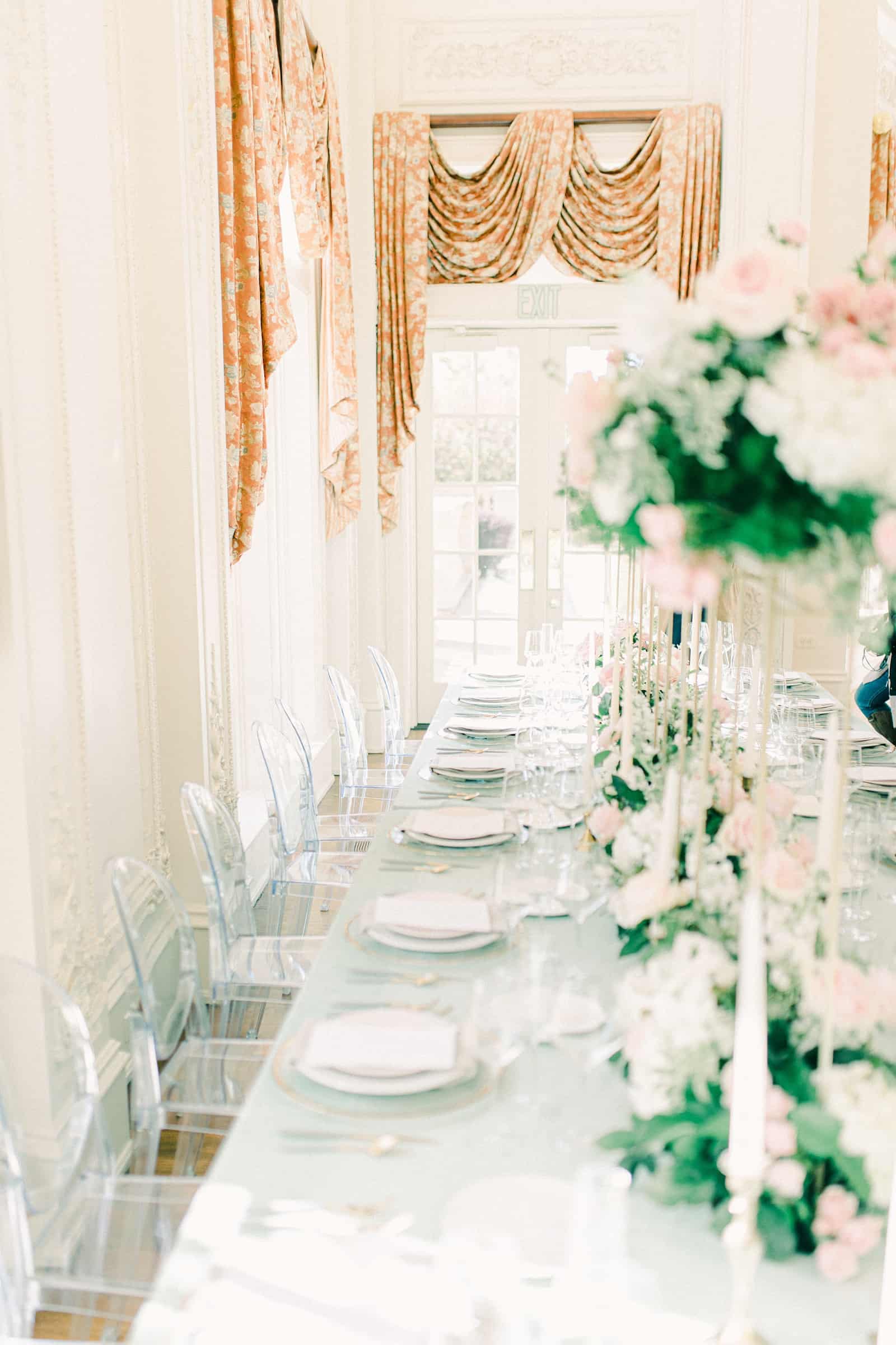 Long wedding dining table with light blue tablecloth, clear dining chairs, tall pink and white flower centerpieces