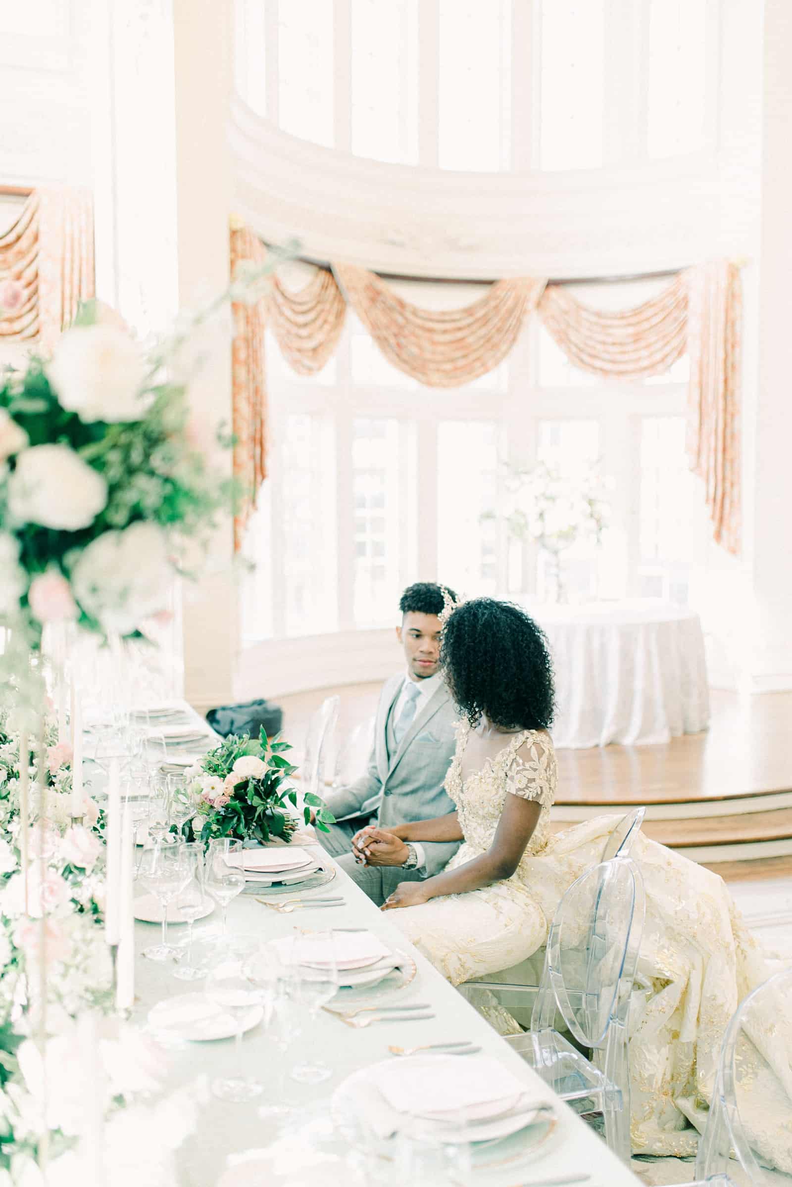 Bride and groom sitting at wedding dining table with light blue tablecloth, clear dining chairs, tall pink and white flower centerpieces, reception decor