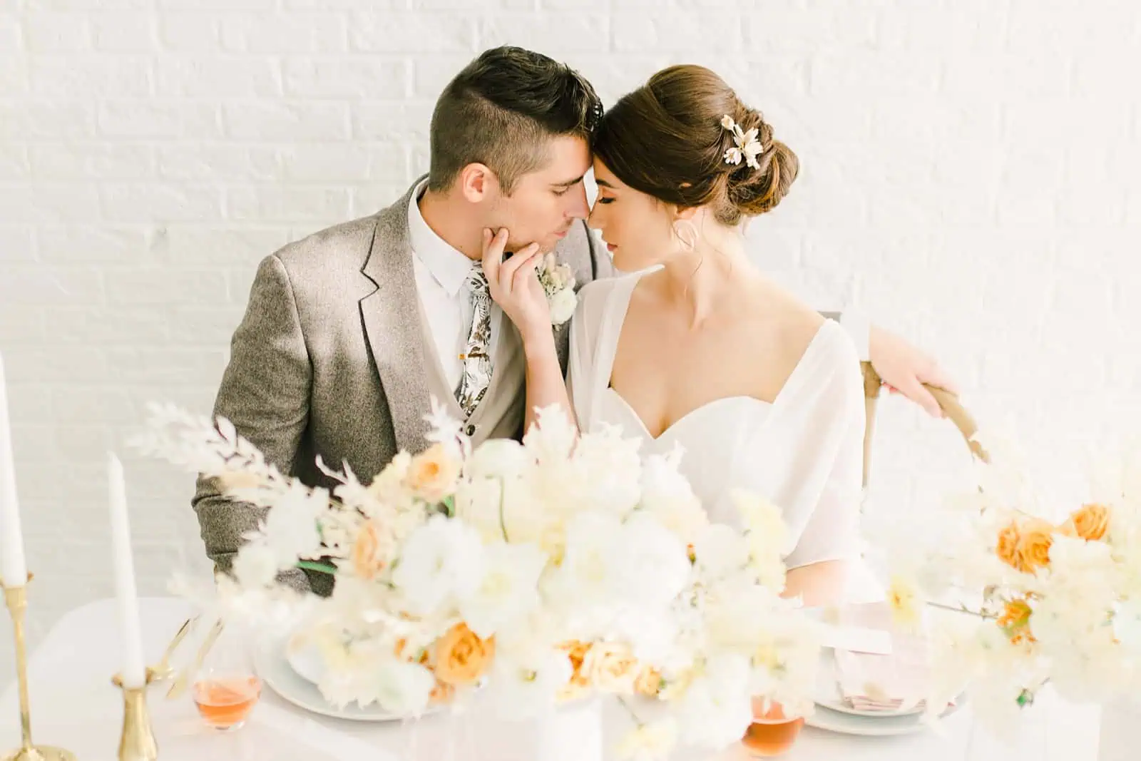 Bride and groom lean close sitting at modern wedding table with white and orange flowers