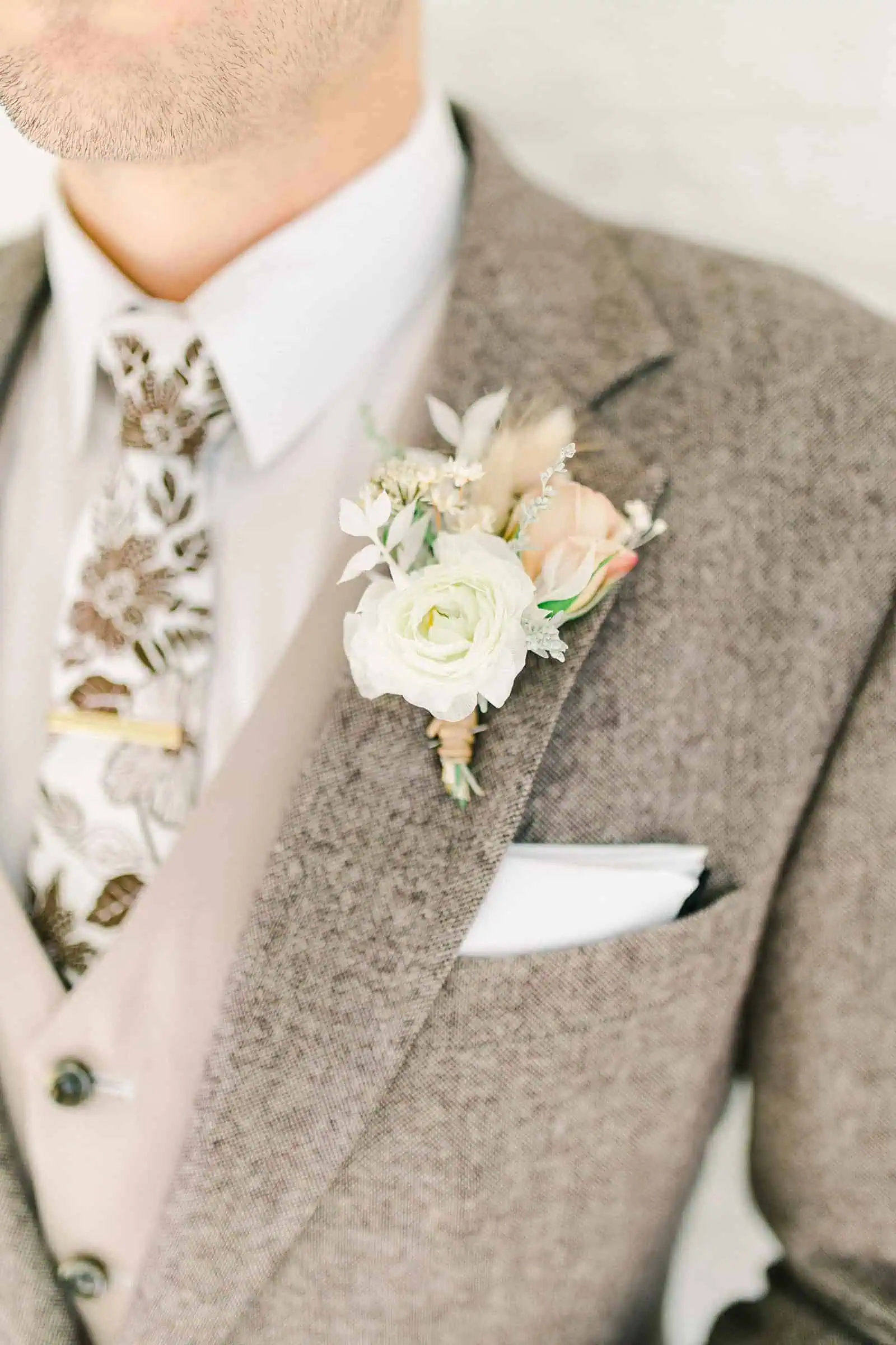 Modern groom wearing oatmeal gray custom three piece suit with fall floral tie and a vest, white neutral boutonniere