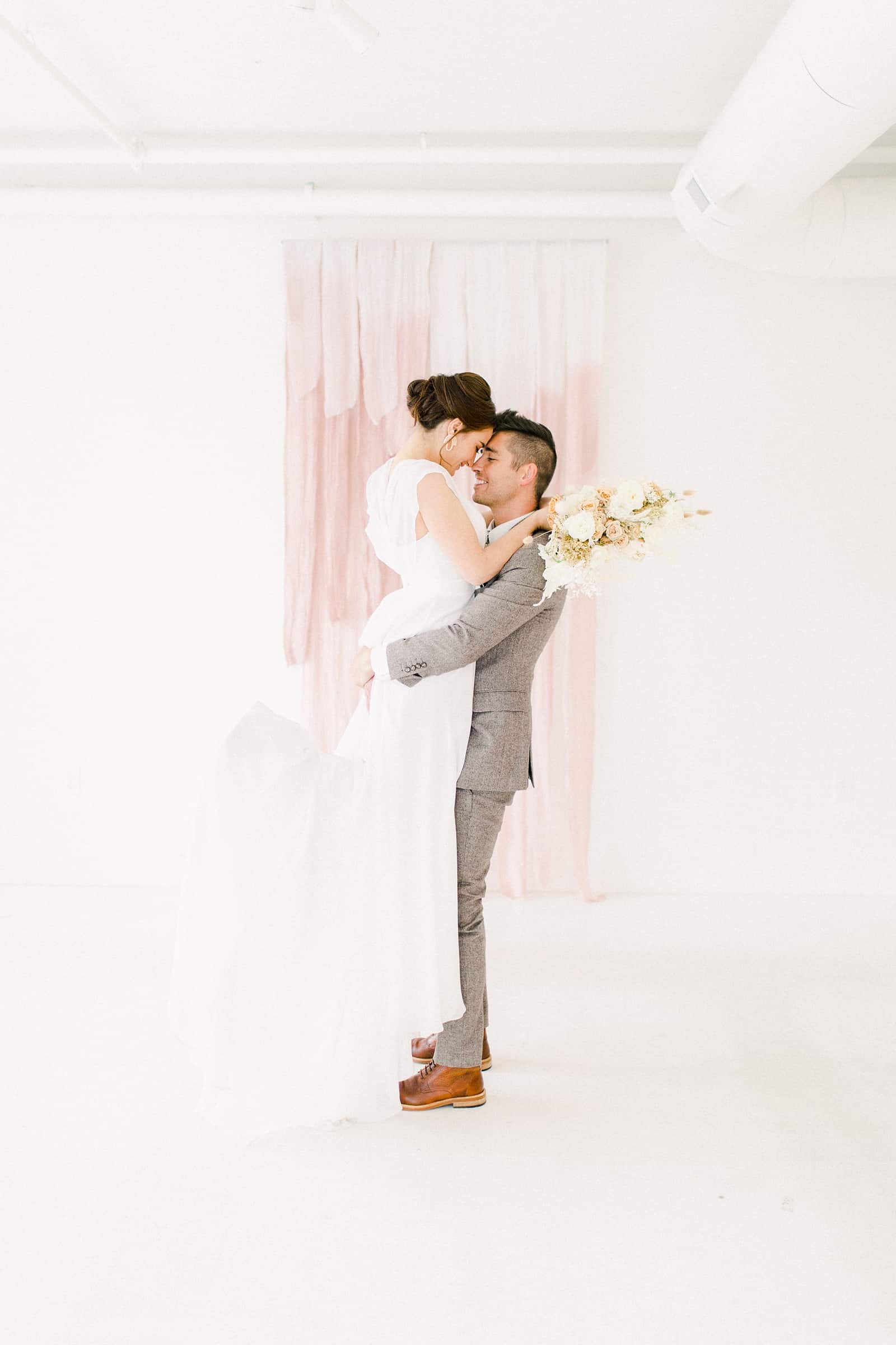 Groom lifts bride after ceremony in front of blush pink painted ombre watercolor backdrop