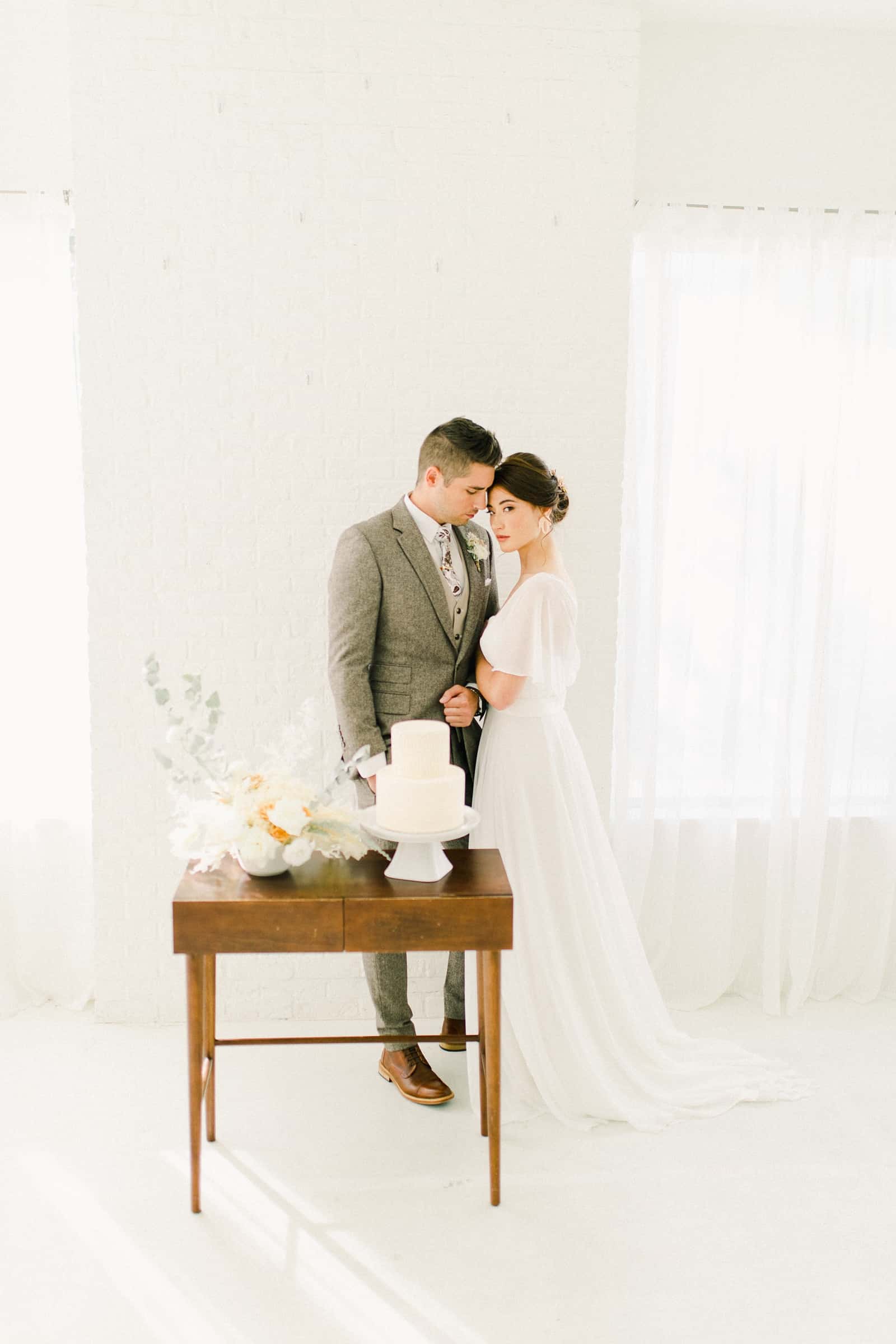 Bride and groom stand in front if Modern wooden wedding cake table with minimalist neutral flowers