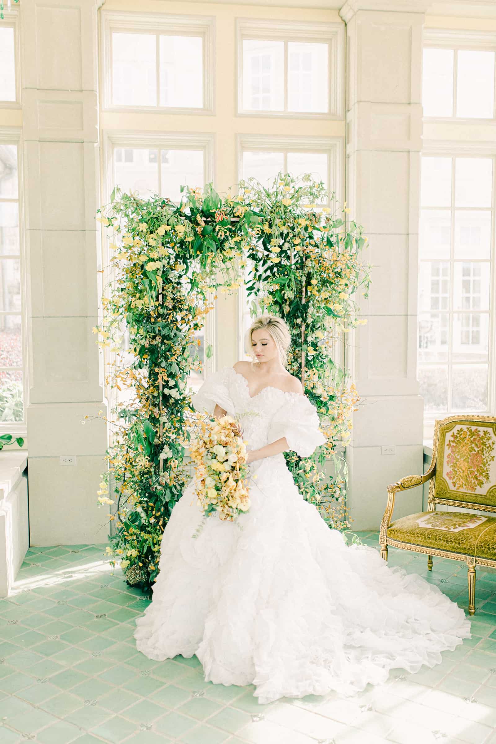 Bride wearing off shoulder ball gown with puff sleeves in front of floral arch