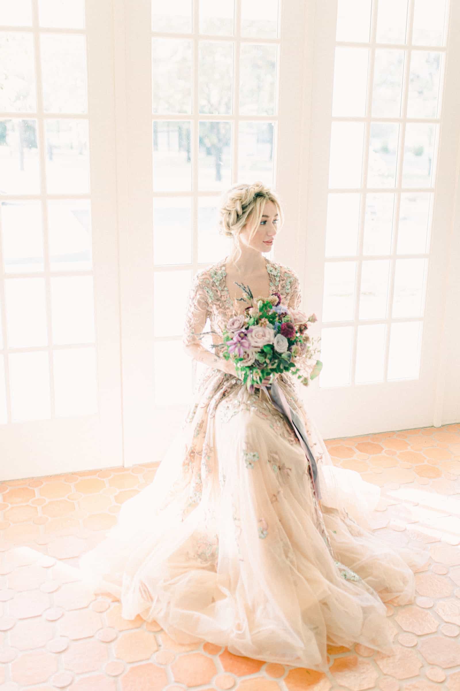 Boho bride in front of large windows with purple flower bouquet