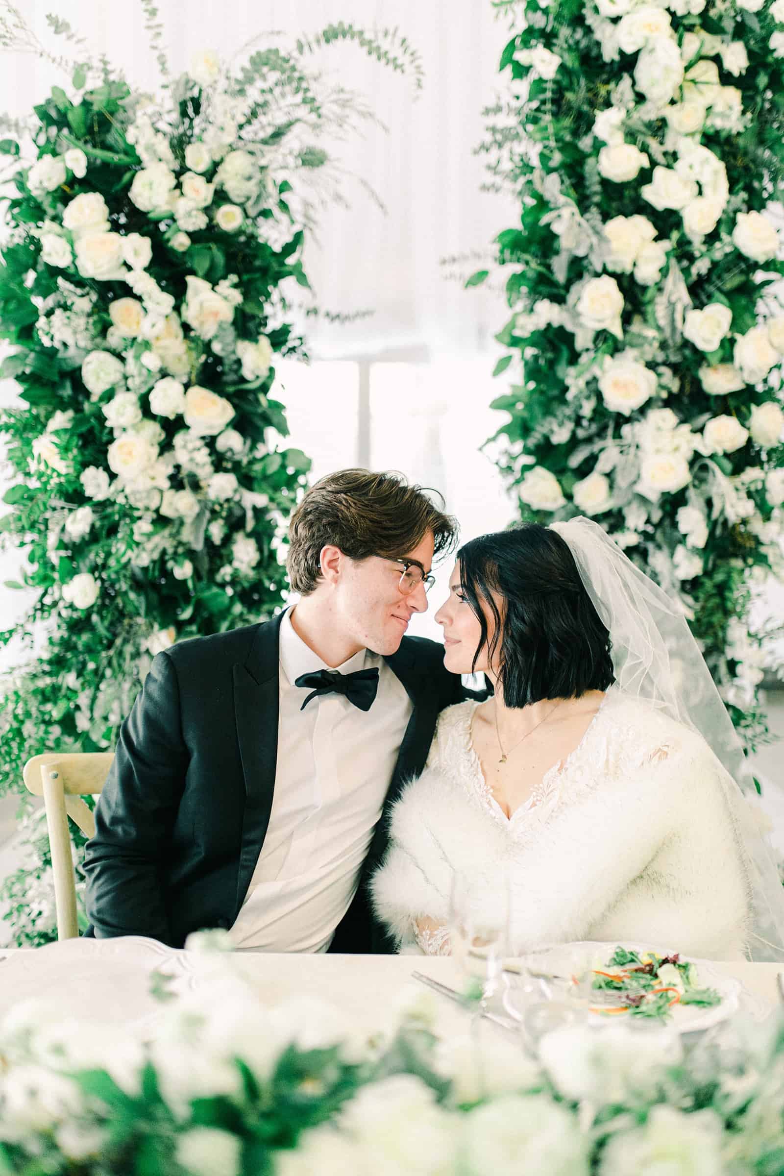 Luxury winter wedding at Willowbridge Estate in Boise, Idaho, bride and groom at sweetheart table with floral arch with white flowers and greenery backdrop