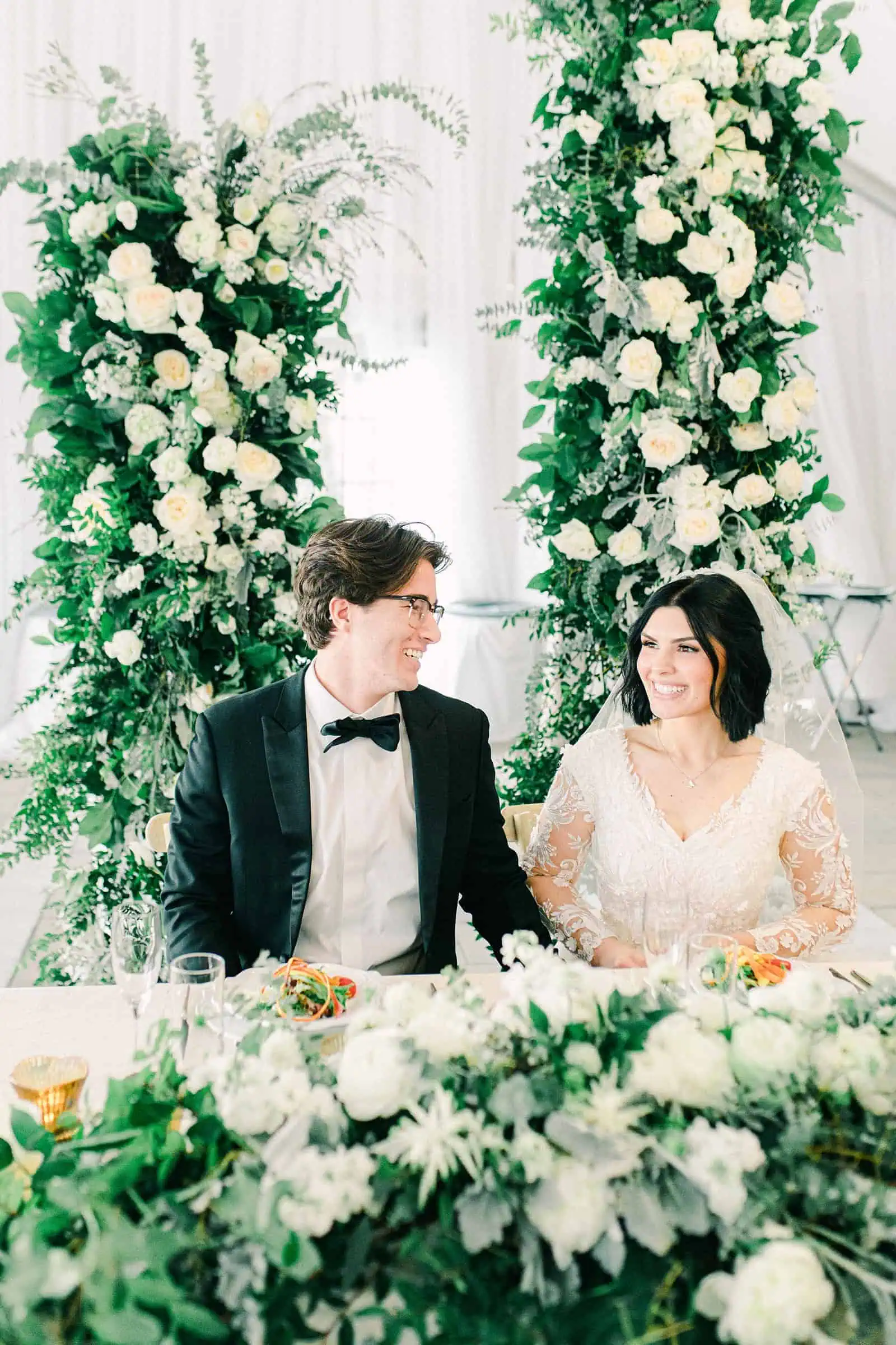 Luxury winter wedding at Willowbridge Estate in Boise, Idaho, bride and groom at sweetheart table with floral arch with white flowers and greenery backdrop