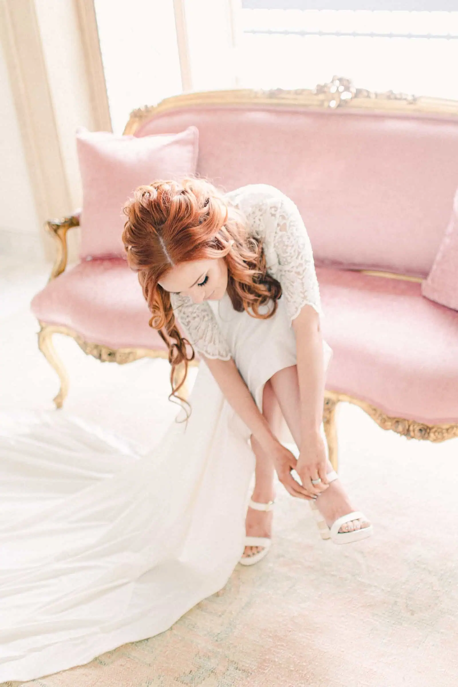 Redhead bride putting on shoes, pink couch in bride's room, bridal suite