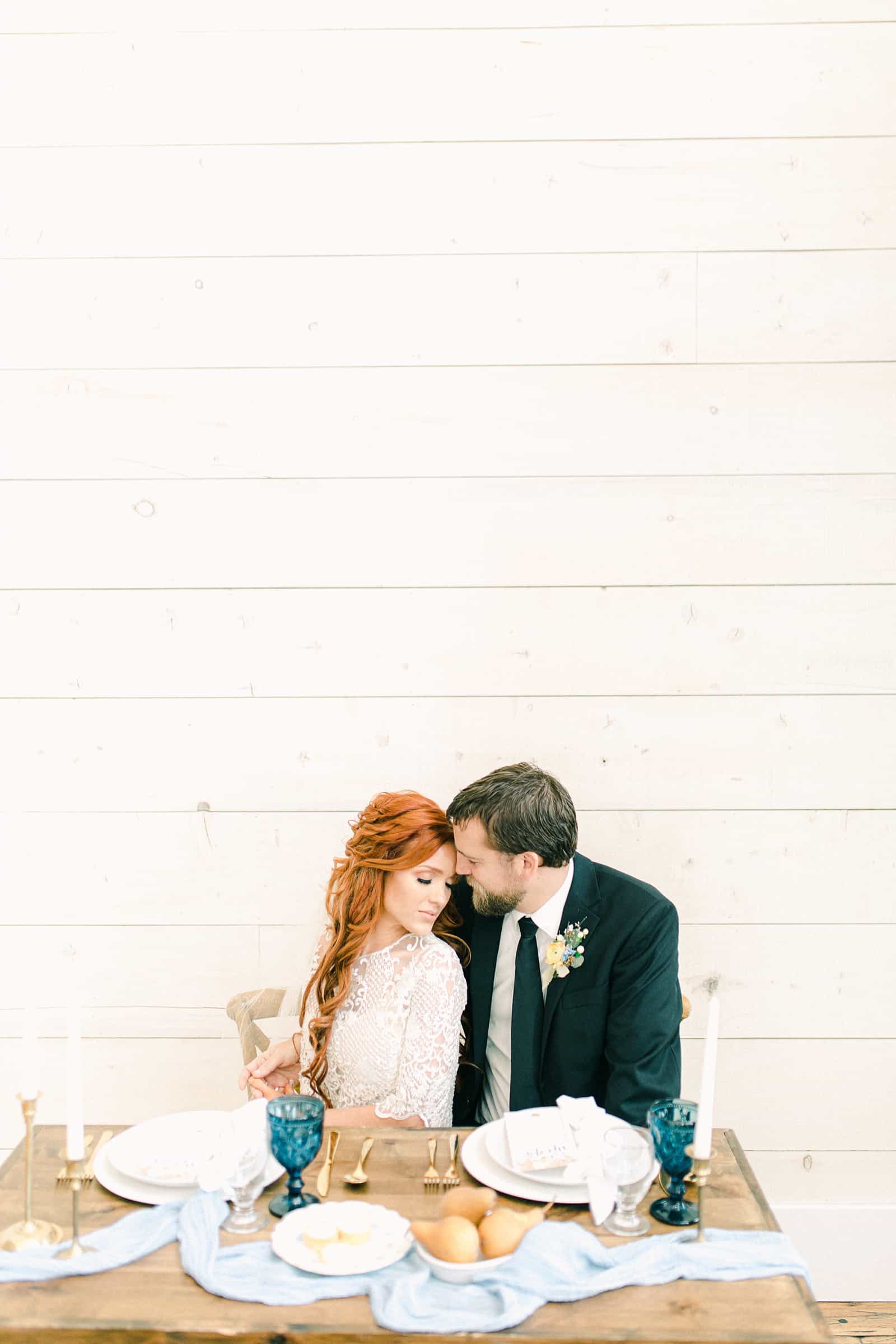 Bride and groom sit at wedding reception table, white shiplap walls, white barn wedding venue