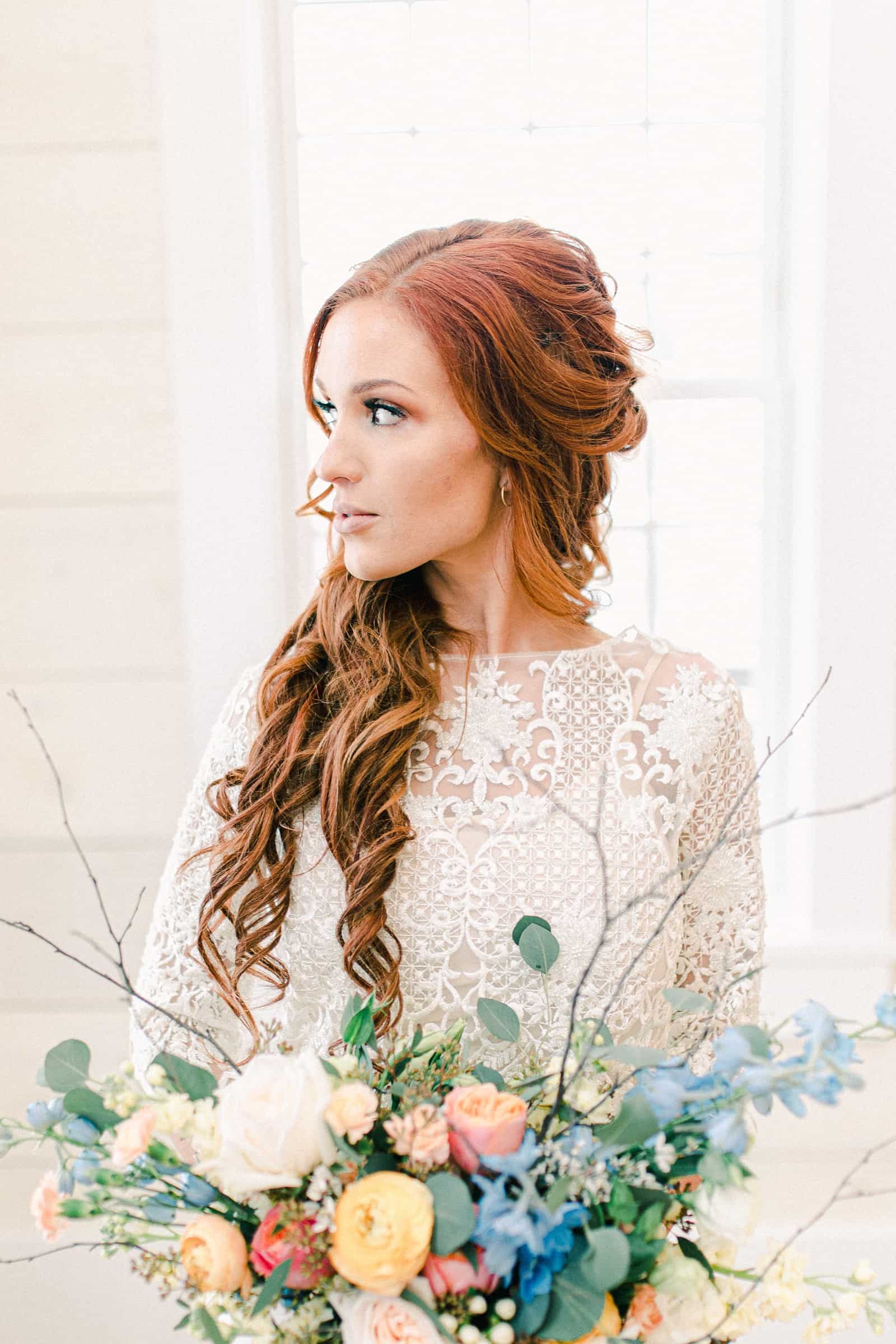 Bride with long lace sleeves holding wedding bouquet with pastel colors, blue, pink, orange, peach flowers, long curly wedding hairstyle redhead, Freckled Fox