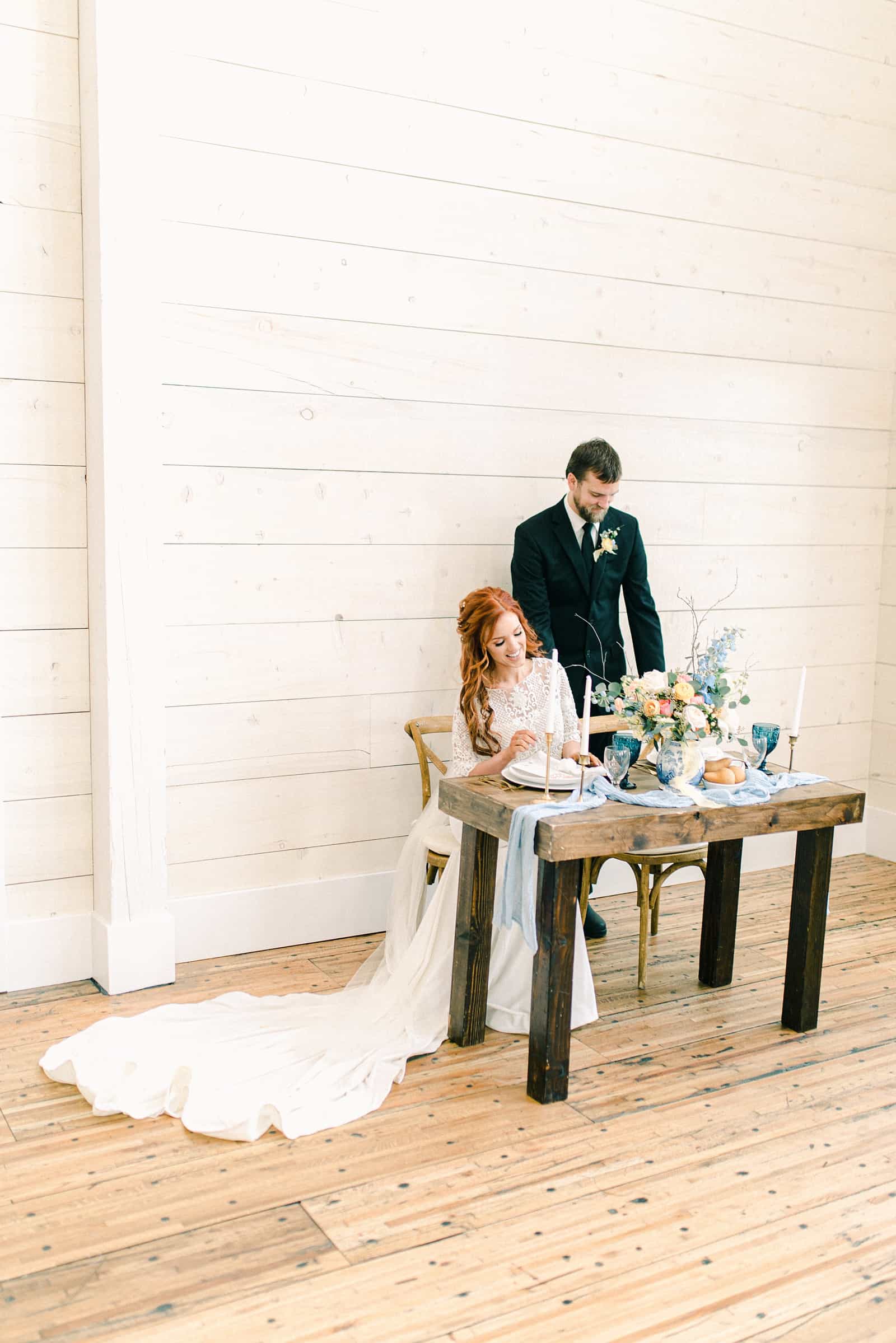 Bride and groom sit at wedding reception table, white shiplap walls, white barn wedding venue