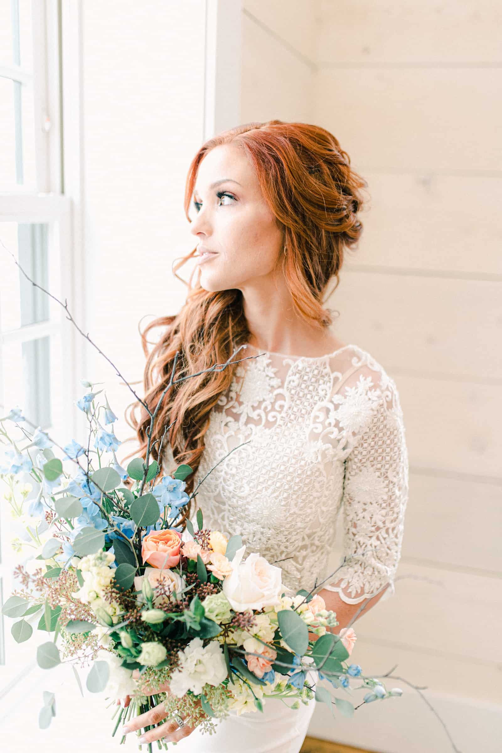 Bride with long lace sleeves looks out window holding wedding bouquet with pastel colors, blue, pink, orange, peach flowers