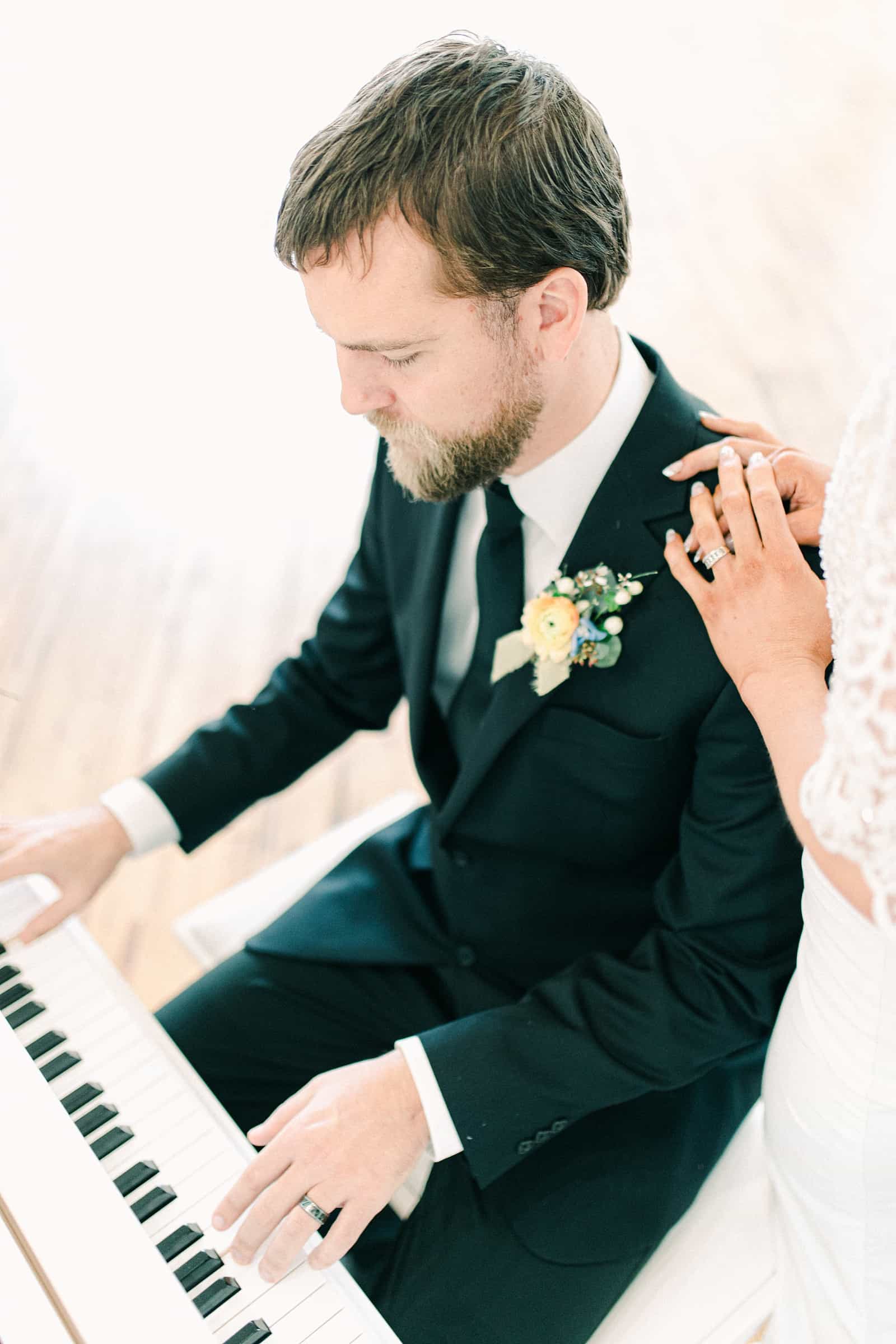 Groom plays white baby grand piano for bride on their wedding day, groom in classic black suit with black tie