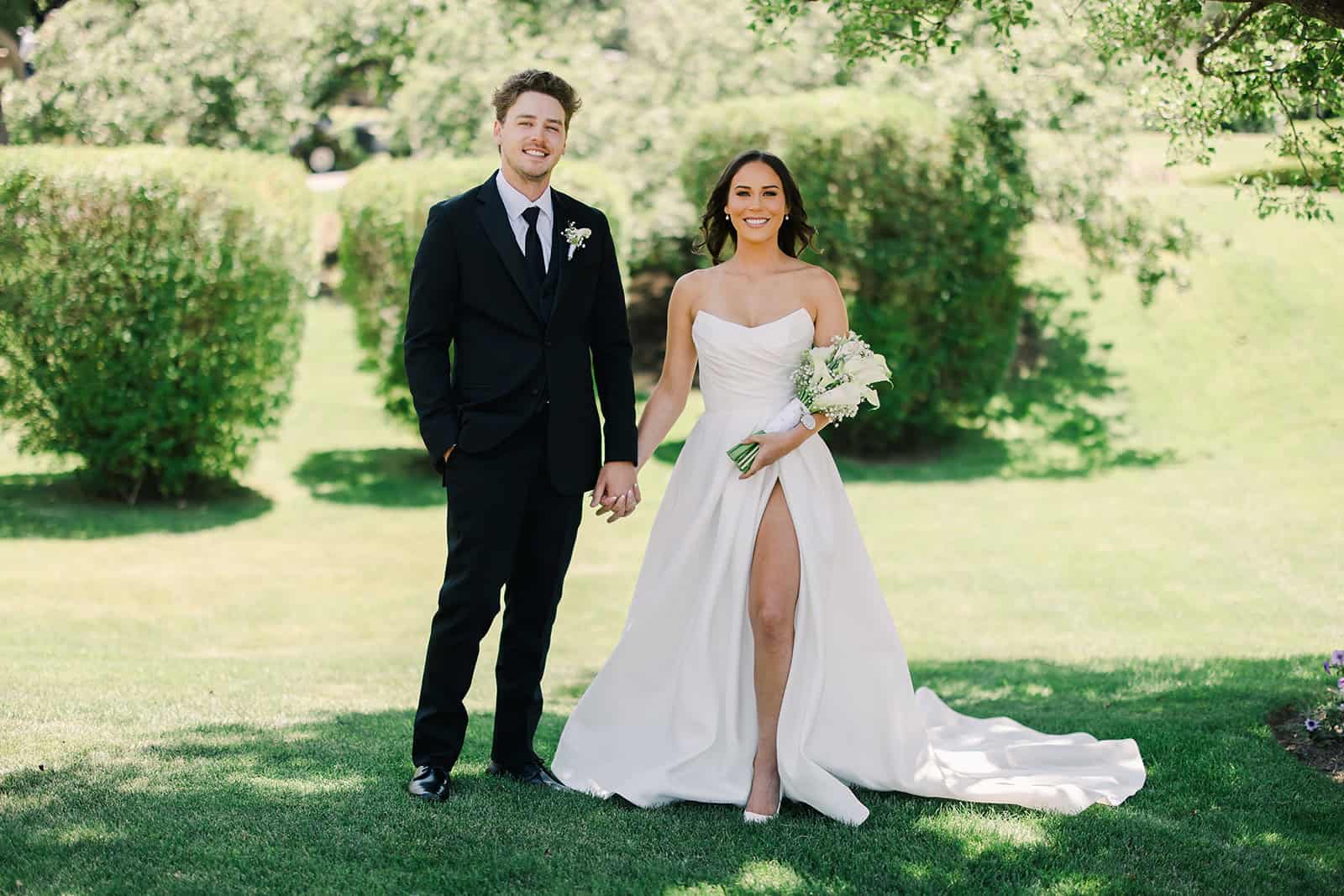 Bride and groom holding hands, smiling, with greenery in the background.