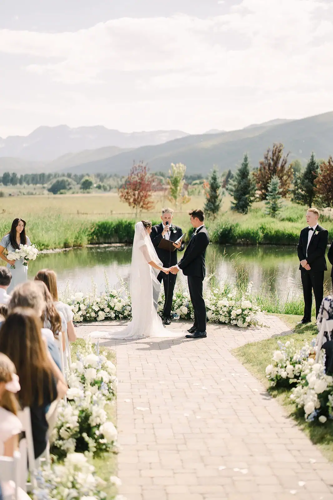 Outdoor wedding ceremony with bride and groom exchanging vows by a pond, surrounded by nature and mo.