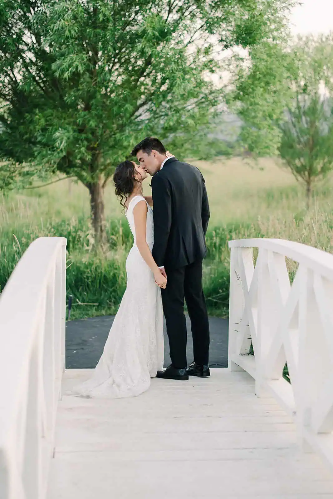 Wedding couple holding hands on a white bridge in a green, natural landscape.
