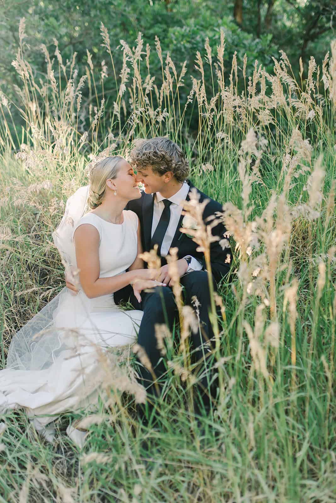 Elegant wedding couple in a lush, green outdoor setting with tall grass and trees.