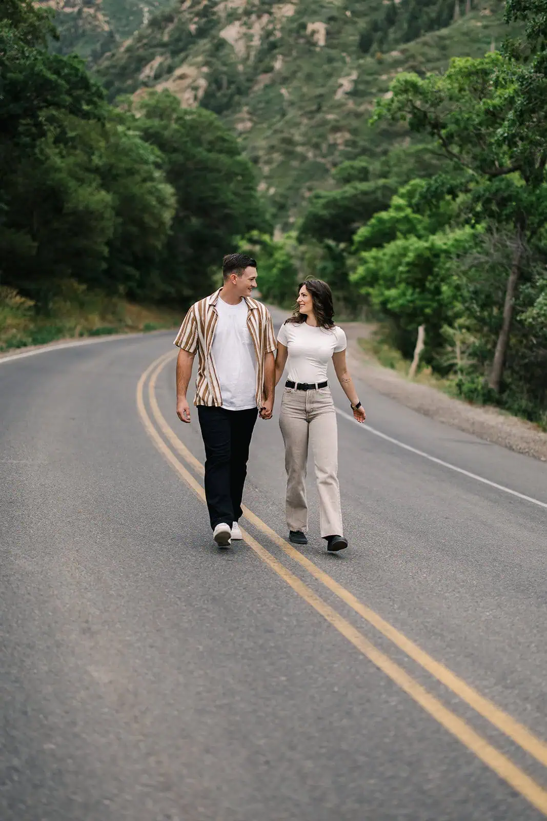 Couple walking hand-in-hand along a winding mountain road surrounded by lush greenery.