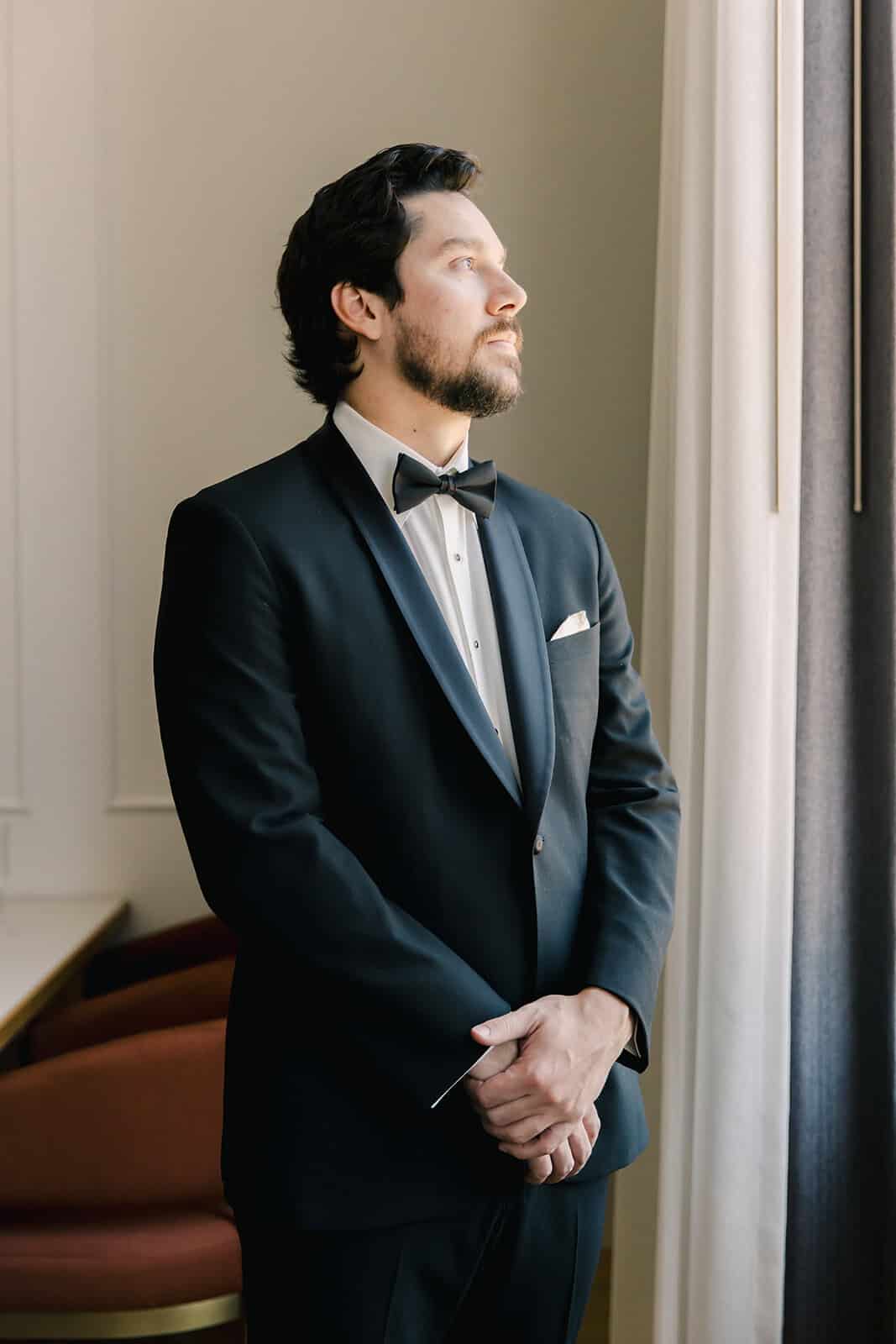 Elegant man in tuxedo gazing out the window, showcasing style and sophistication.