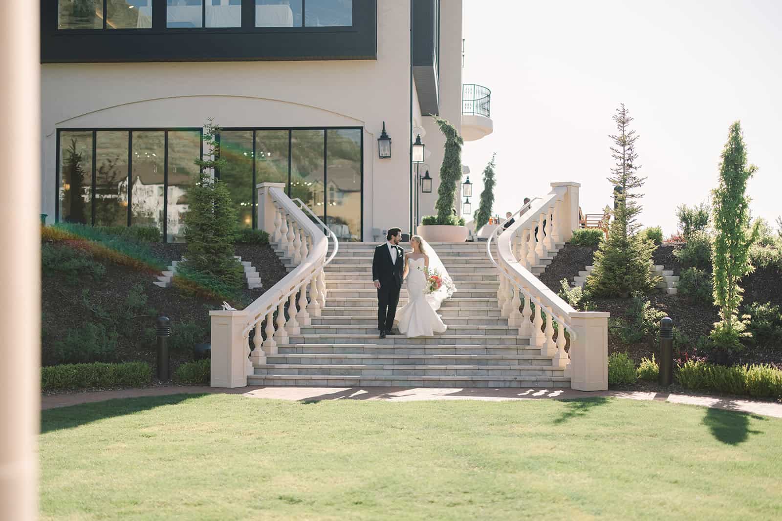 Elegant wedding photo of bride and groom on grand staircase with lush landscaping.