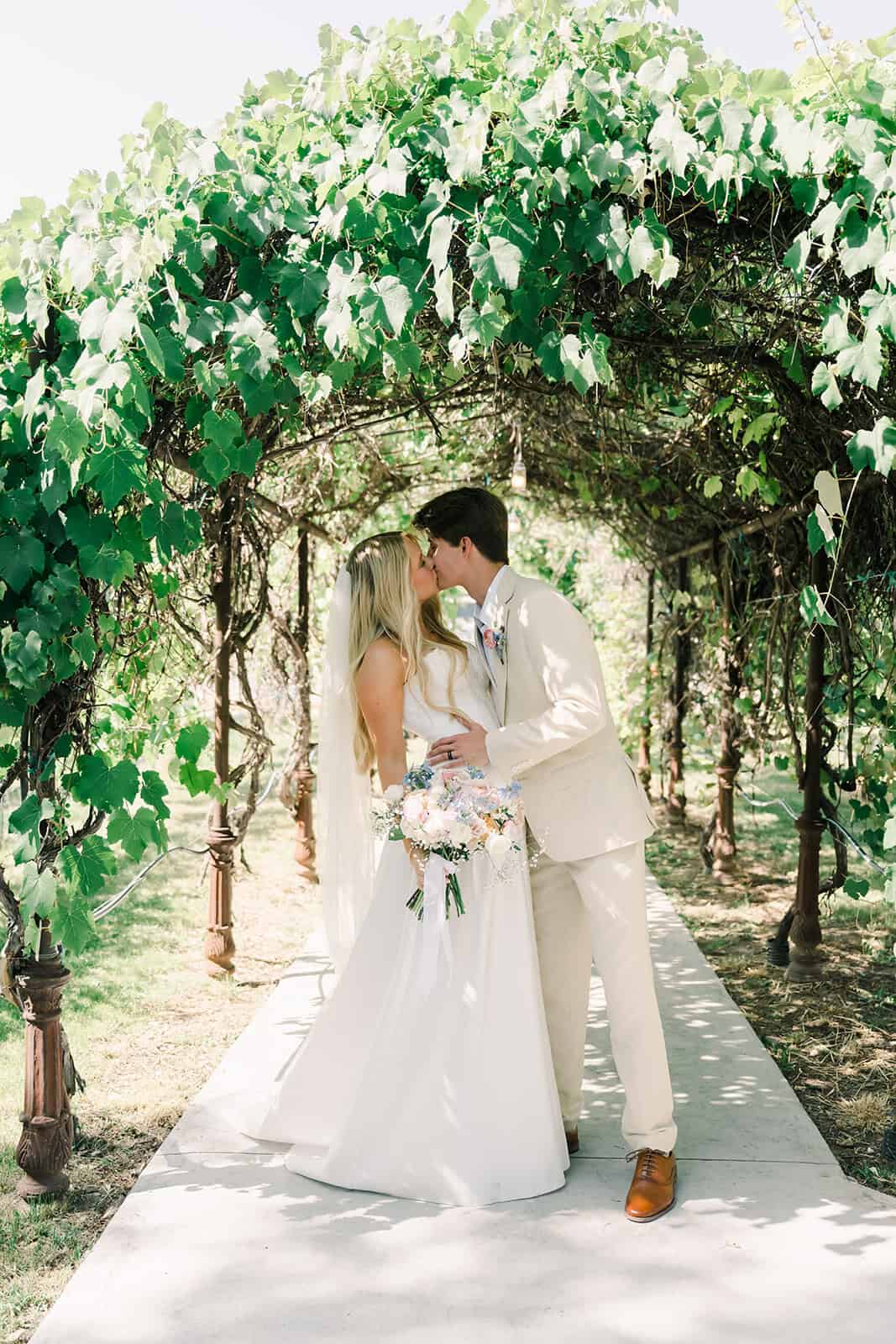 Romantic wedding couple sharing a kiss beneath a lush green archway.